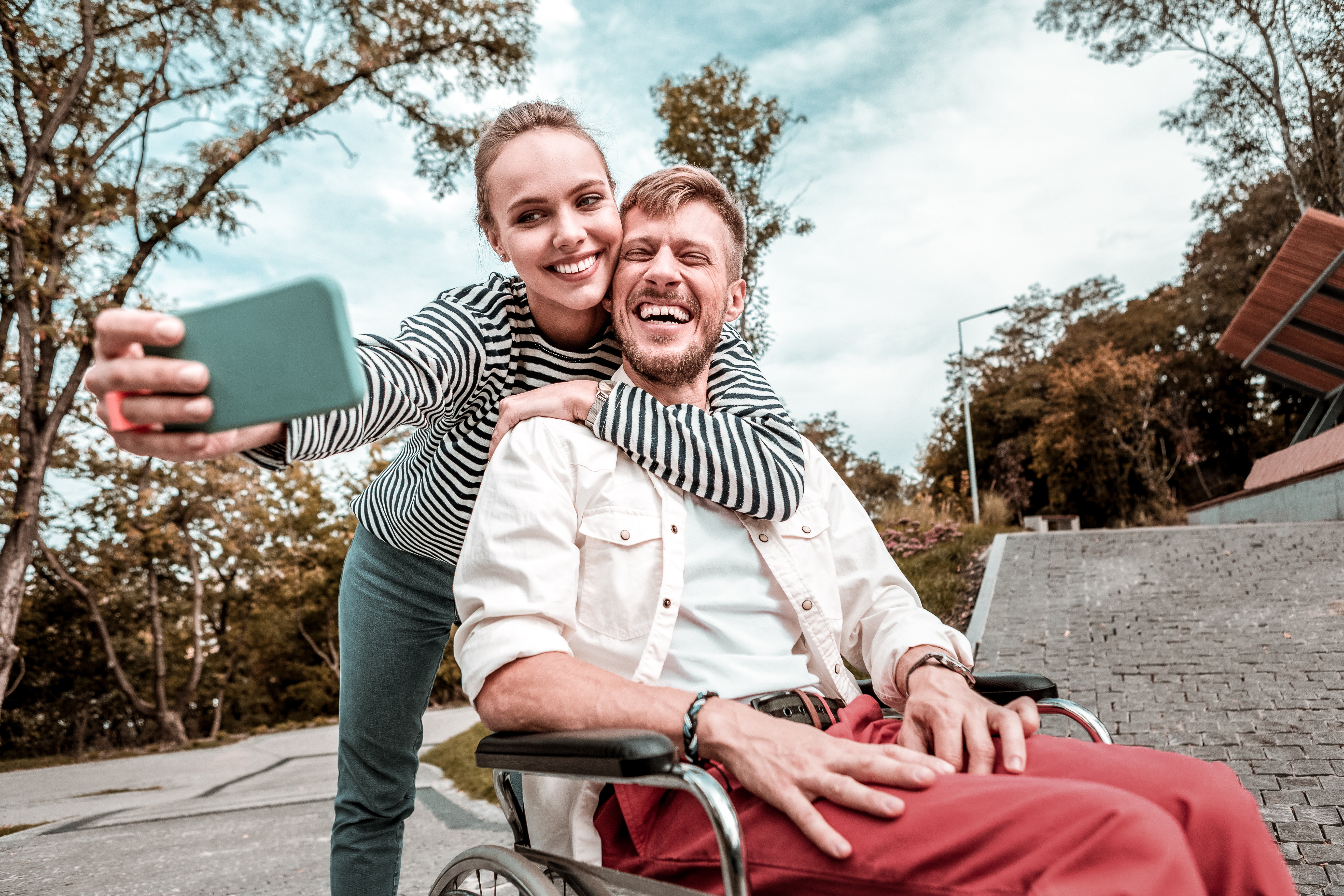 a woman holds up a mobile phone taking a selfie of herself and the man she is with in a wheelchair.