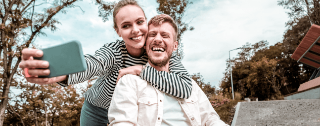a woman holds up a mobile phone taking a selfie of herself and the man she is with in a wheelchair.