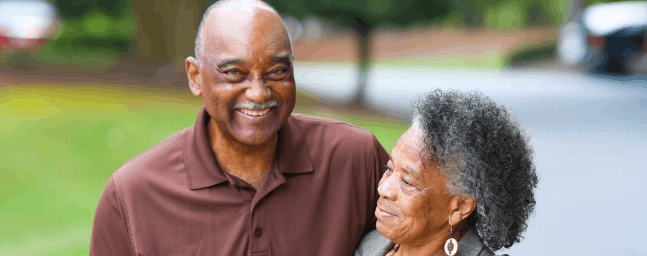 older couple, man wears a brown top and has his arm around a woman who wears an orange top and grey jacket. Both are looking at each other.