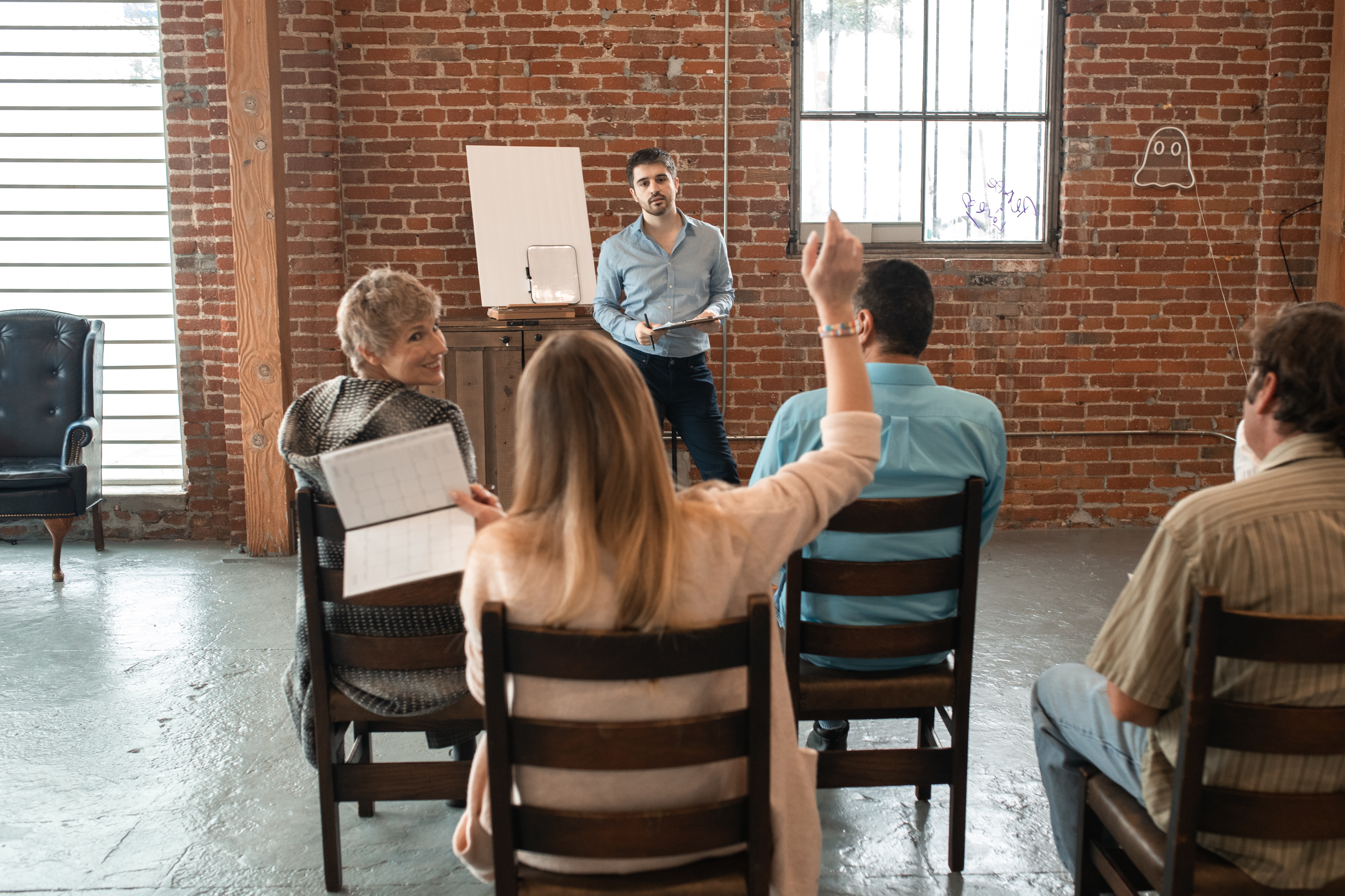 Photo shows a group of people at a meeting in chairs facing a man with a whiteboard who is presenting to them. There is a woman with her hand up waiting to as a question